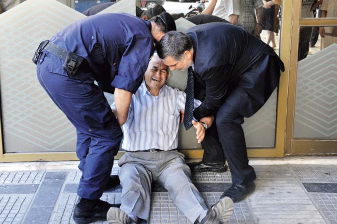 A crying elderly Greek man is assisted by an employee and a policeman outside a national bank as pensioners queue to get their pensions, with a limit of 120 euros