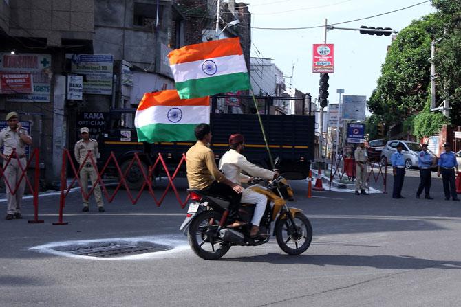 Men on a motorbike carrying Indian national flags ride past security personnel guarding a street as the country celebrates it's 73rd Independence Day in Jammu