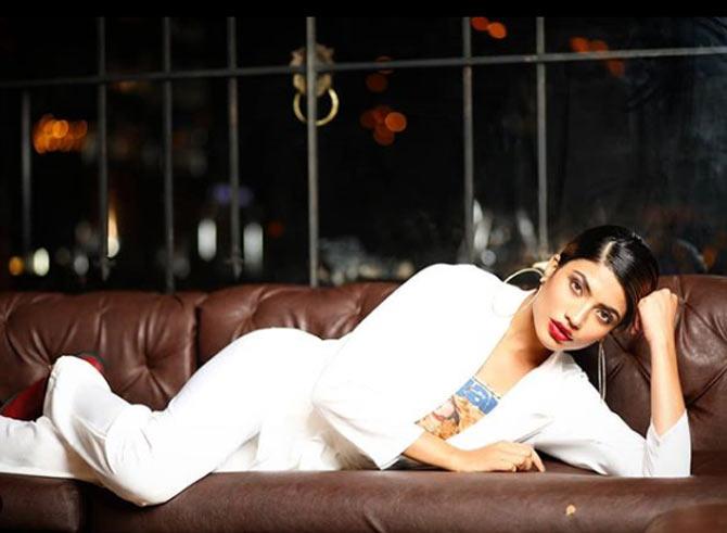 In this photo, Urvi Shetty poses on a couch in a white suit and trousers along with a blue-printed top