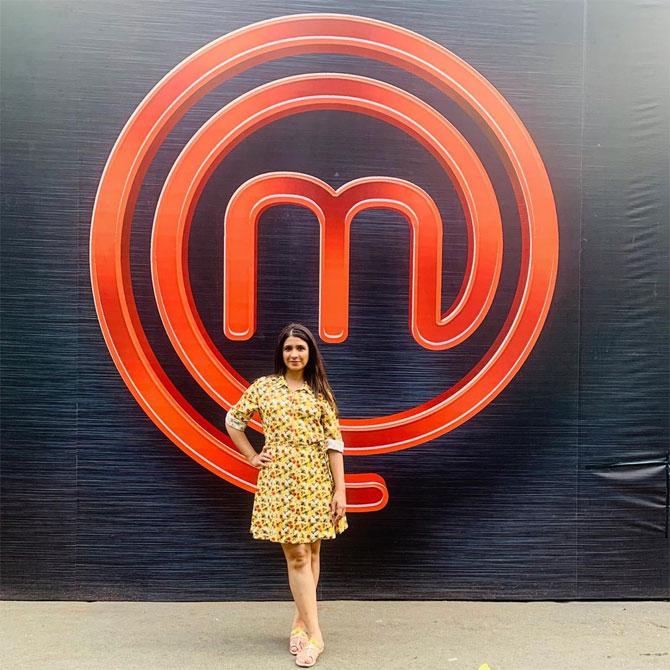 In picture: Akanksha poses in front of the Masterchef India logo in a yellow printed dress and her hair left open.