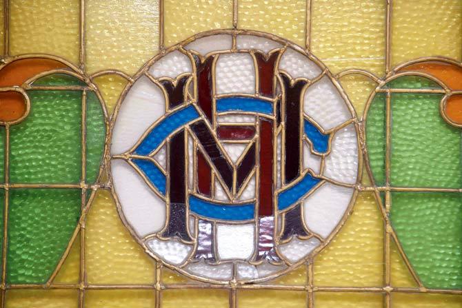 A stained glass window with the family crest initials of the Colahs who lived at 8, Club Road. From their balcony they clearly saw the Bombay Central Station clock across open maidan stretches and could hear the zoo animals of Rani Baug