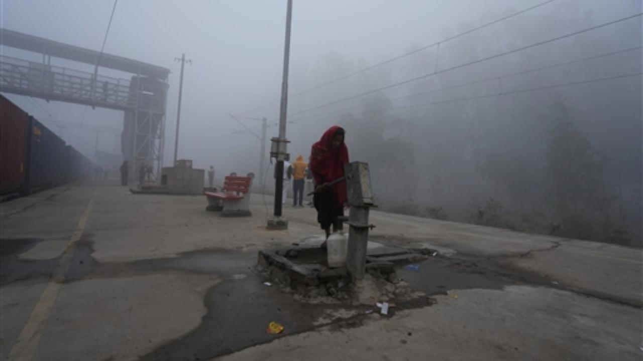 A woman collects water from a hand pump amid dense fog on a cold winter morning, at the Maripat Station in Greater Noida
