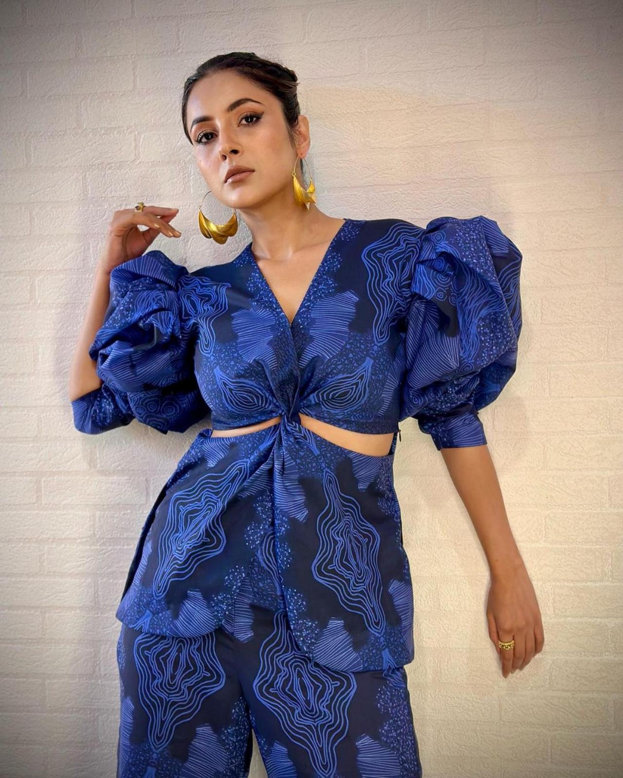 Shehnaaz truly 'blue' us away with her navy blue printed jumpsuit accentuated with dramatic puff sleeves. Her apparent love for statement earrings slips in again as she opts for geometric golden earrings. 