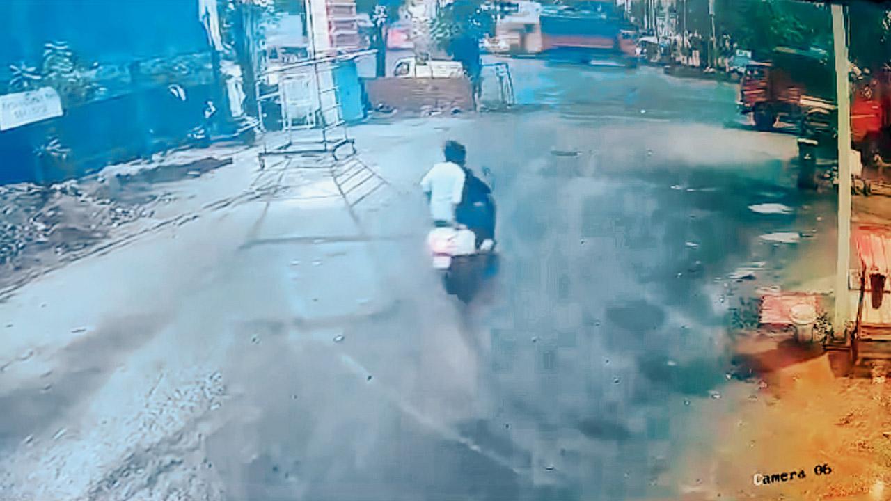 Two men on a scooter approach a busy, unmanned intersection. They are not wearing helmets