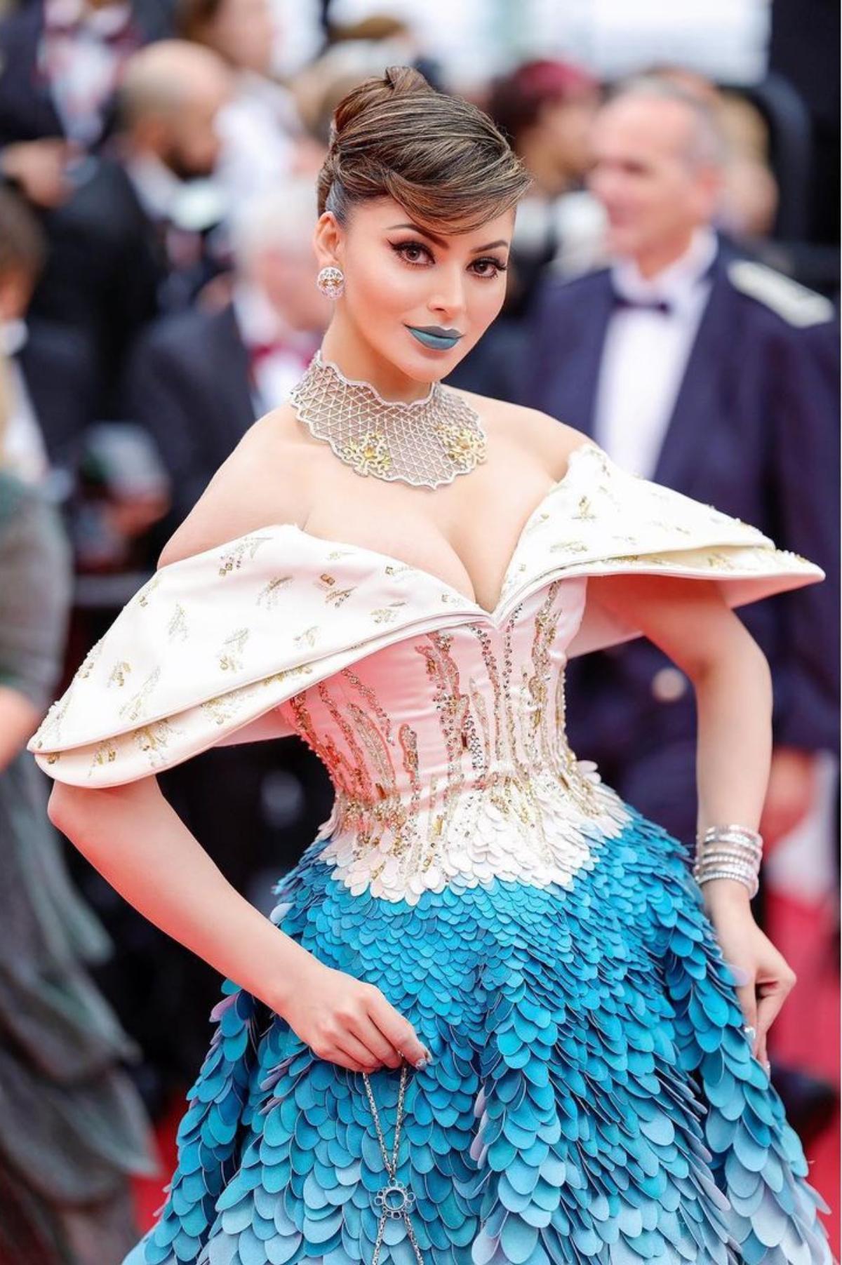 Urvashi Rautela who was heavily trolled for wearing a Cartier crocodile necklace with a pink tulle gown, once again, caught everyone's attention as she arrived wearing a voluminous blue and white off-shoulder ruffled ombre gown and painted her lips blue to complete her red carpet look on day 3. 