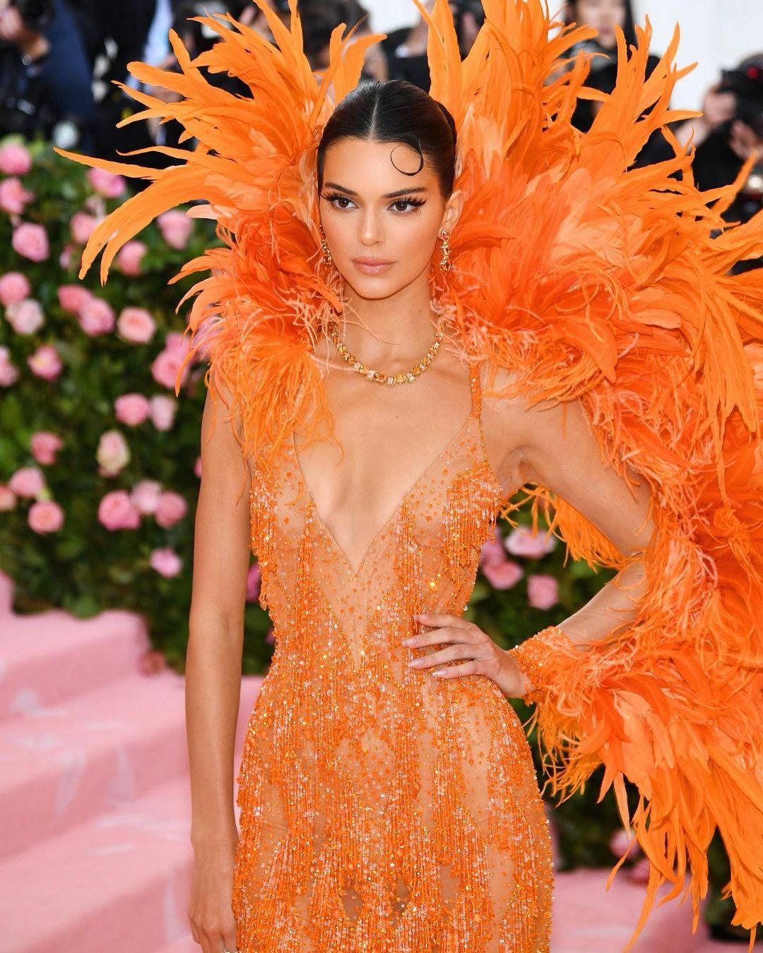 A Kendall Jenner fashion gallery starting with this look is a must. Kendall Jenner wore this huge orange feathered Versace gown with her hair slicked back in a flapper-esque style for the Met Gala in in 2019. This look has gone down in history as one of her best