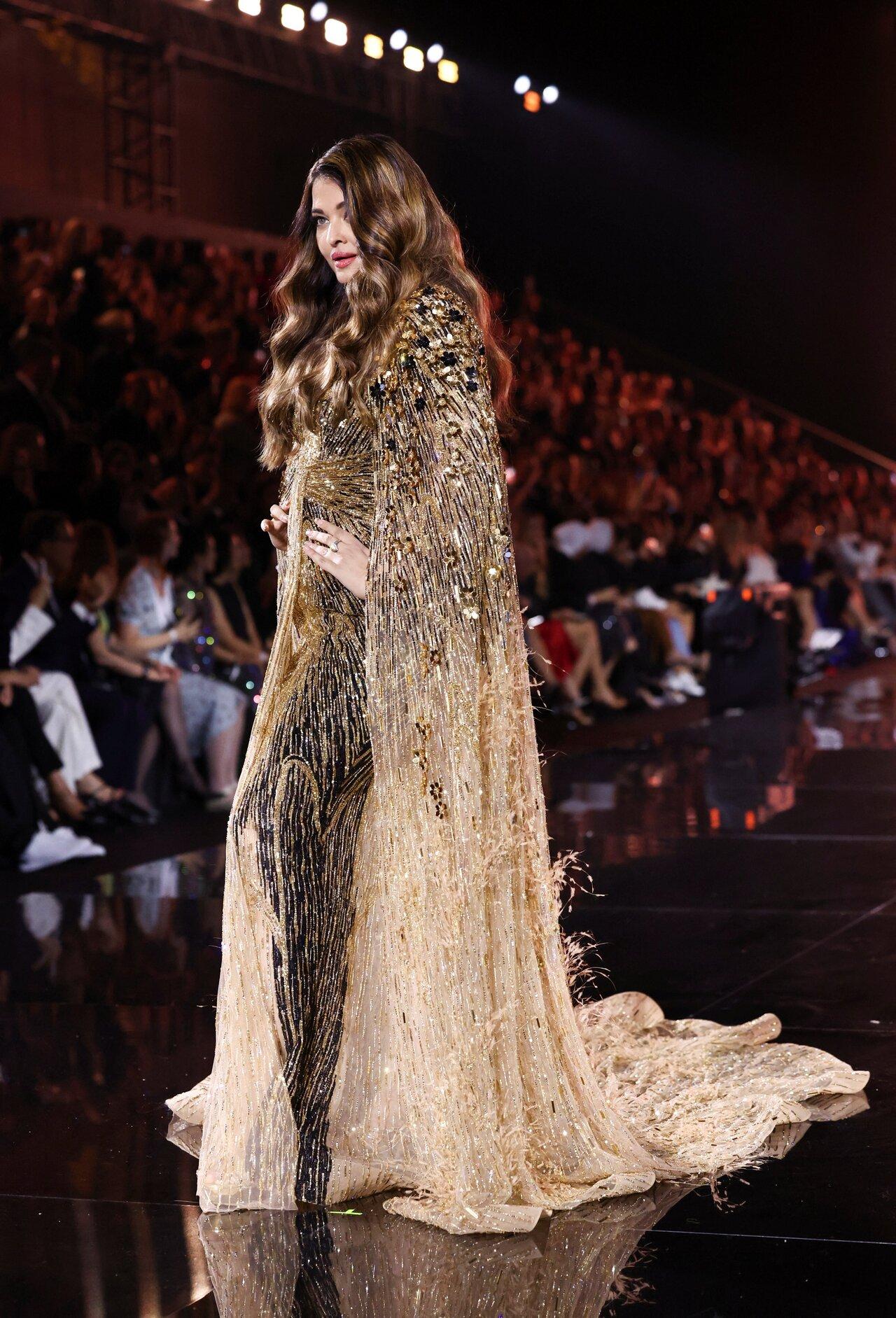Aishwarya's golden gown features sparkling gold sequin embellishments, beaded embroidery, a bodycon silhouette highlighting her curves, a gathered design on the front, a see-through cape attached on the back, and a floor-sweeping length forming a train