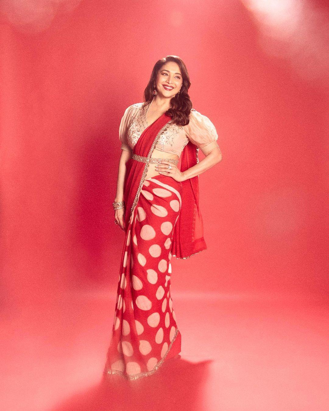 Madhuri Dixit's outfit for Diwali is a stunning red and gold polka dot saree that beautifully captures the essence of elegance and is a perfect pick for the occasion. The breezy drape of the saree strikes a harmonious balance between chic and stylish