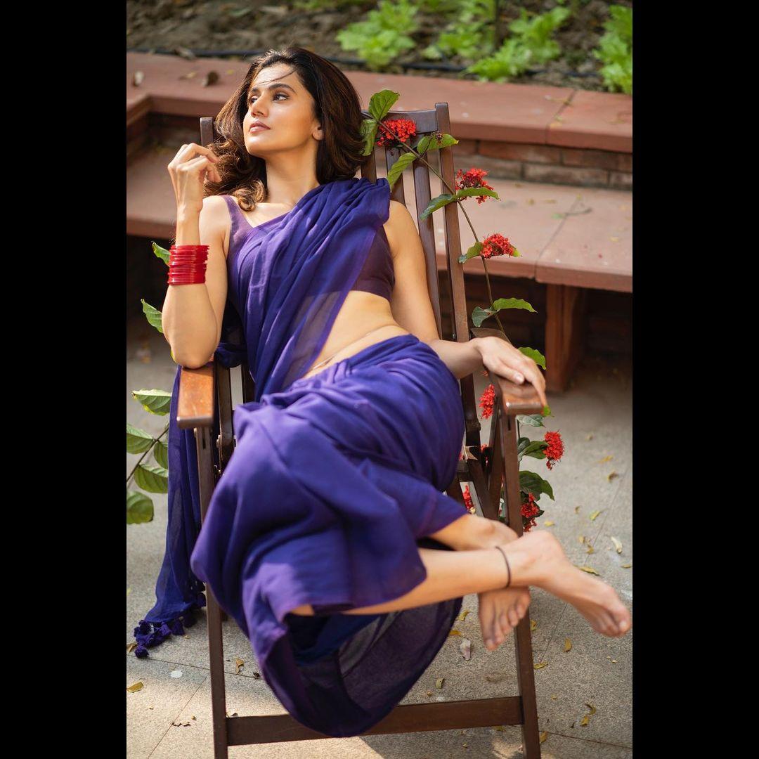 Channeling 90s vibes, Taapsee poses on a wooden chair in this purple saree, paired with some red bangles