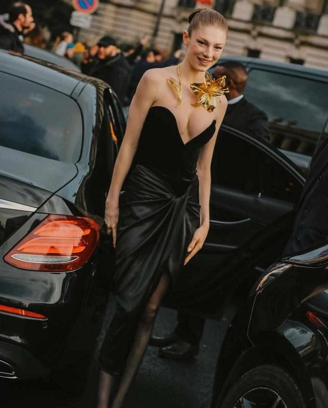 Hunter Schafer wore a black strapless velvet midi dress that had a gathered waist, a side slit, and a flowy satin finish – typical Schiaparelli elegance. Staying true to their signature style, a touch of gold was added to the dress. The strapless neckline, featuring a deep v-neck, served as the ideal backdrop for the open-front golden floral necklace.