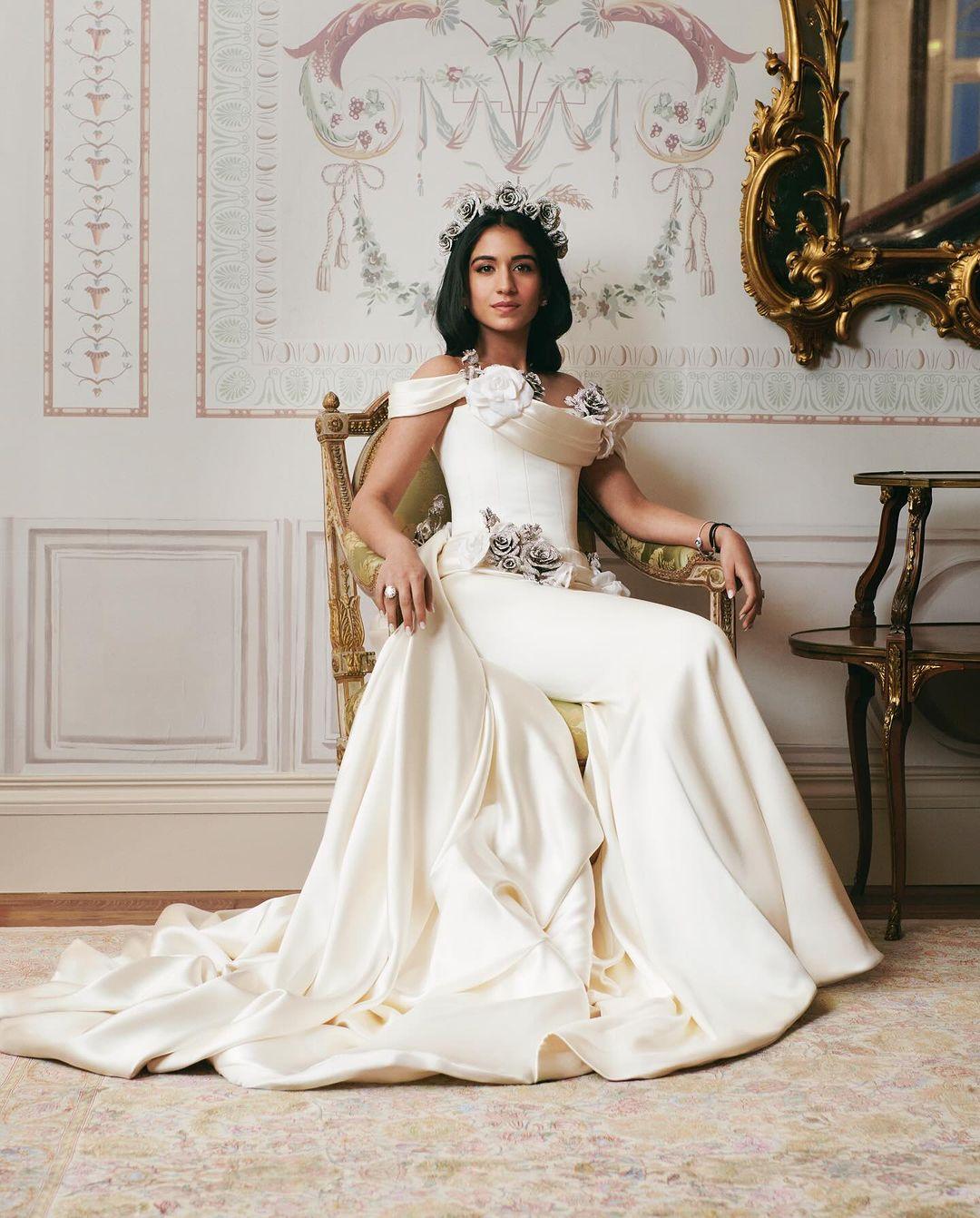 Radhika Merchant wore a beautiful gown with a white silk overskirt decorated with crystal roses. The dress had an off-shoulder neckline, a crystal rose headband, and a long train. She completed her look with diamond jewellery, including earrings, a big solitaire ring, and a delicate bracelet. She styled her hair in loose waves, parted in the centre with a flower crown atop.