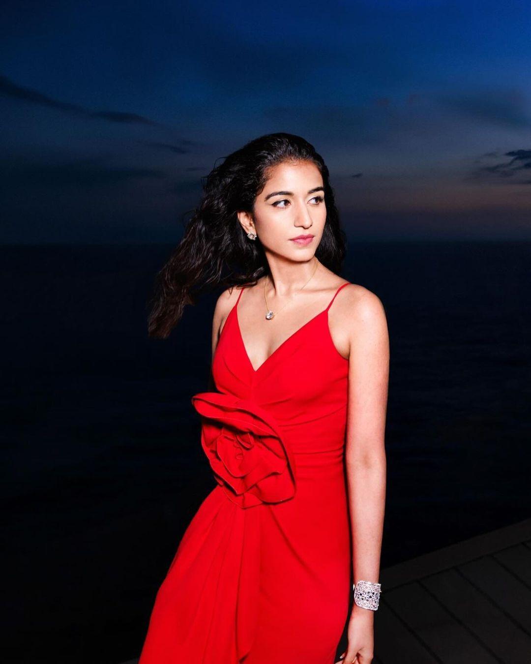 The dress has a deep V neckline, a floral detail on the front, pleats cascading down, a figure-hugging fit, and a floor-length hem. Shaleena styled Radhika's look with a matching red scarf, a diamond wrist cuff, a diamond pendant on a delicate chain, diamond stud earrings, and statement rings.
