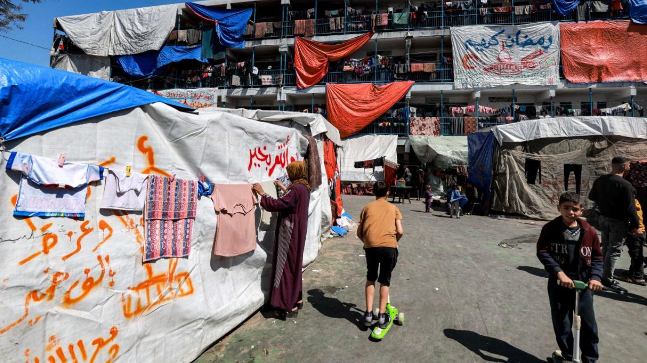 A woman hangs out laundered clothes to dry outside a tent inscribed with greeting messages for the Muslim holy month of Ramadan, at a camp sheltering displaced Palestinians erected in a school run by the United Nations Relief and Works Agency for Palestine Refugees (UNRWA) in Rafah in the southern Gaza Strip