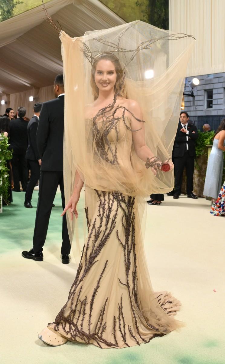 Lana Del Ray made a stunning comeback to the Met Gala tonight, looking like a mystical forest spirit. She wore a gorgeous beige gown designed by Alexander McQueen, with Sean McGirr's touch evident in the intricate thorny brown twigs wrapping around her body. 