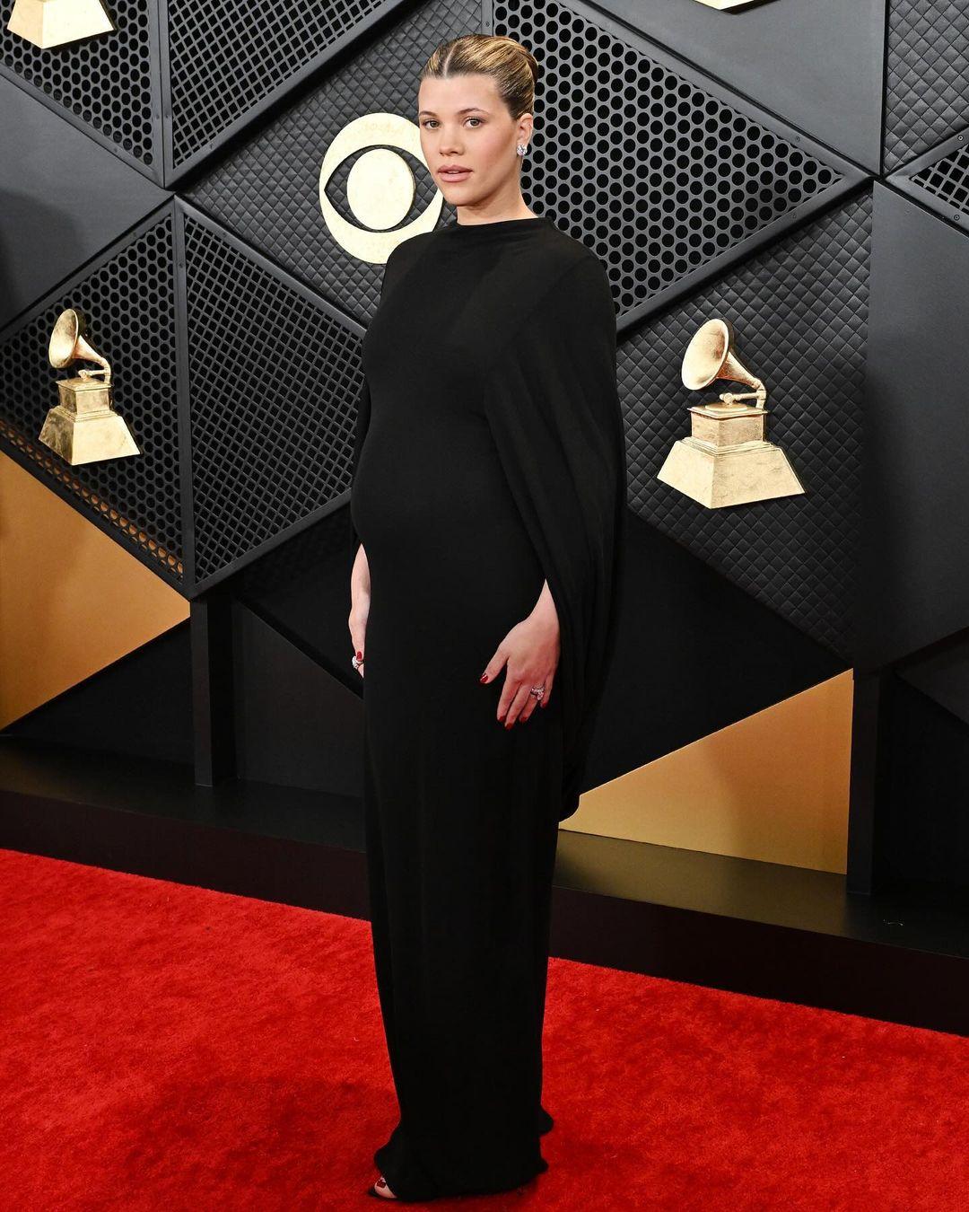 For the Grammys, Sofia Richie wore a timeless black dress with elegant cape sleeves designed by Saint Laurent, complemented by large diamond stud earrings and rings. She opted for a natural makeup look, a red manicure, and styled her hair in a neat ballerina bun.