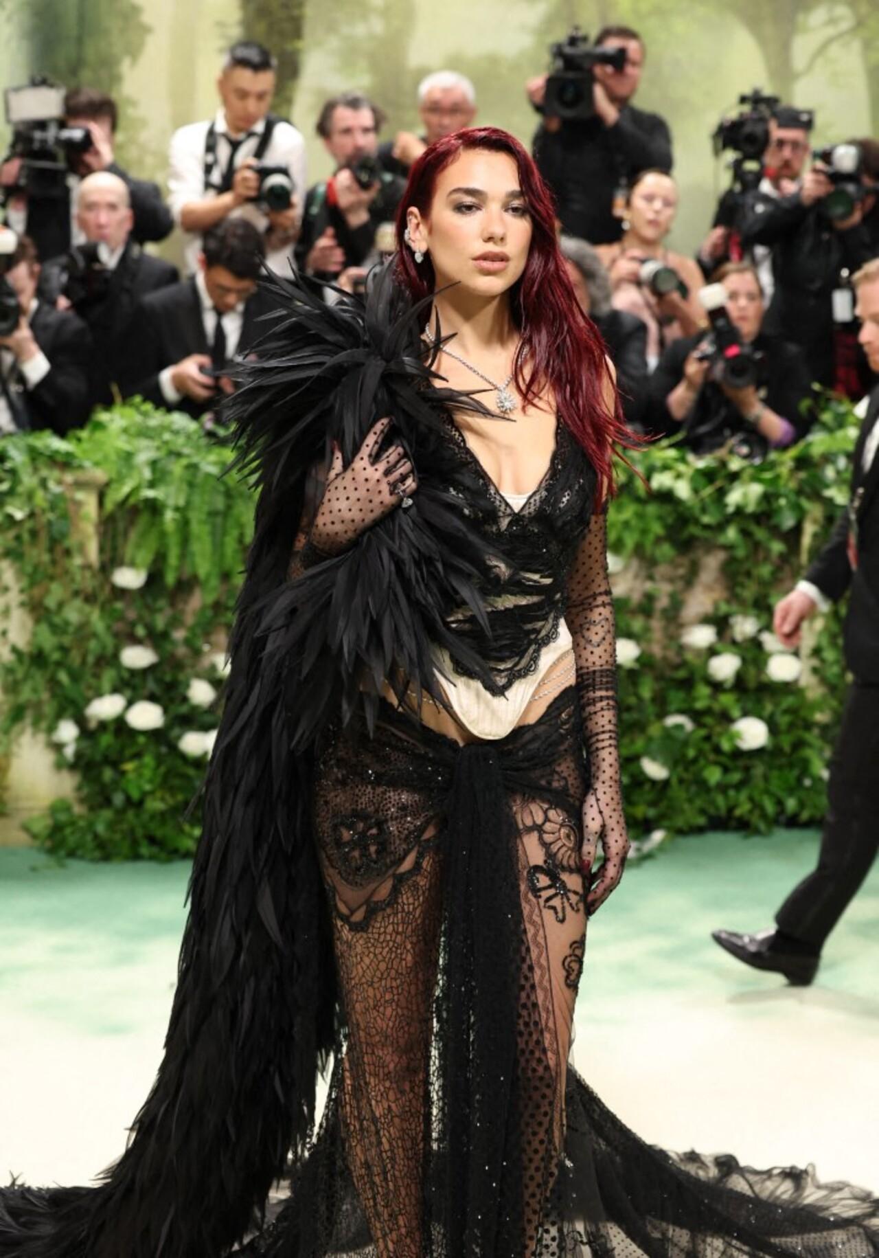 Singer Dua Lipa served an all-black look wearing a skin-bearing lace skirt and corset set that featured an array of intricate details sure to inspire some double takes. To top off the lace black-and-cream look, the Radical Optimism artist accessorized with a black feather boa draped across her shoulder, sheer gloves, a diamond necklace, and a diamond body chain that draped across her low waist. 