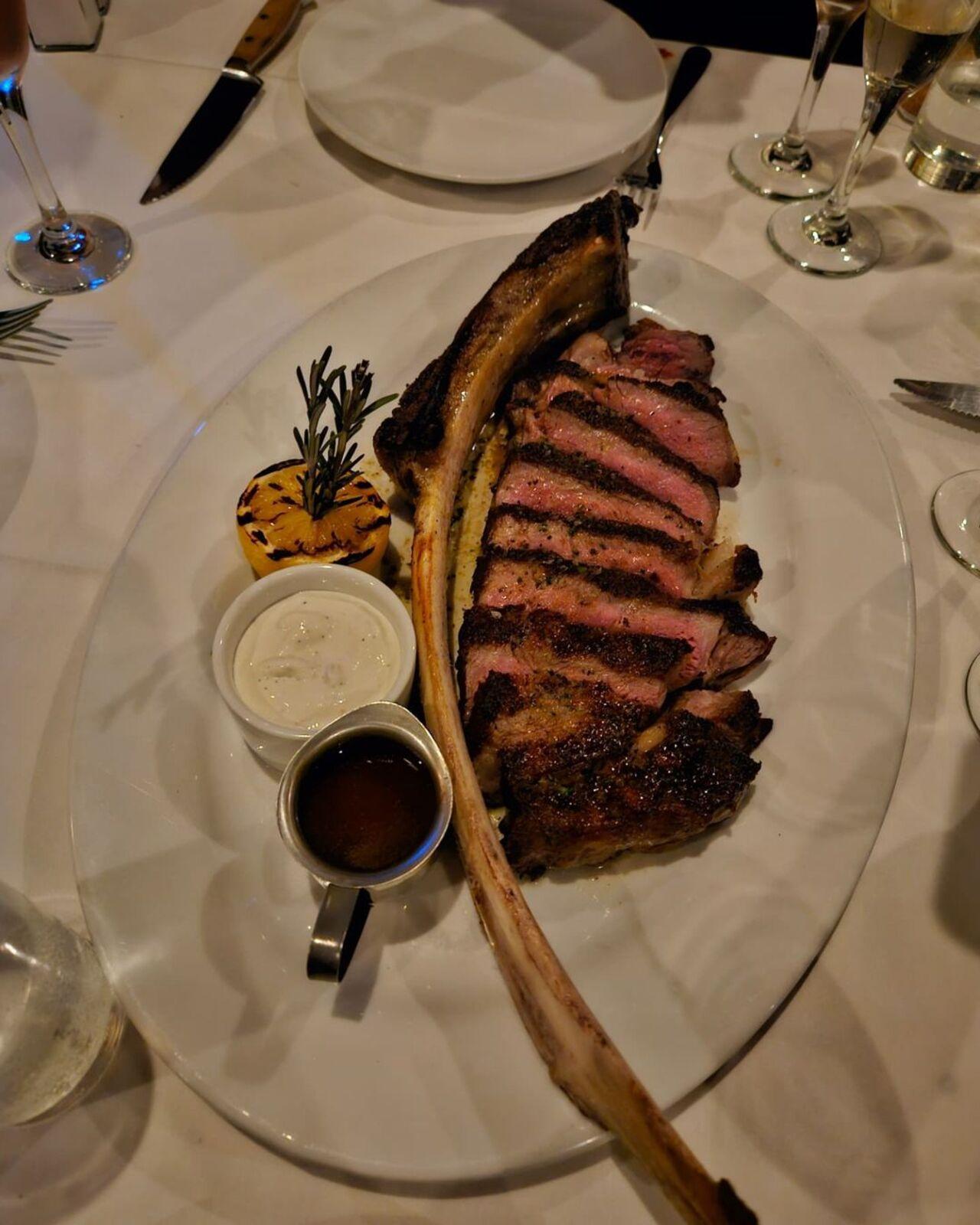 J-hope enjoyed some tomahawk steak with a charred crust. It was served with a charred lemon, a sprig of rosemary for garnish, and two sauces — possibly horseradish or aioli, and au jus or a steak sauce. 