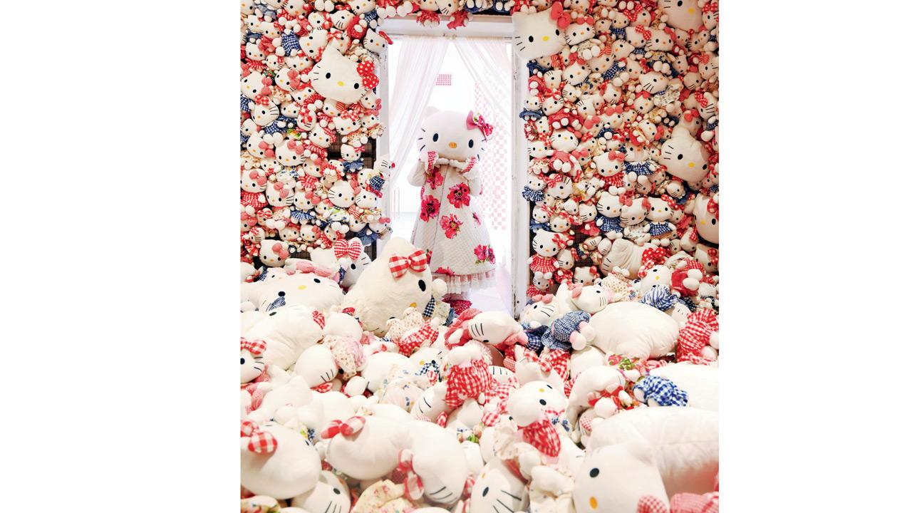 Hello Kitty becomes a textile story by Péro: a real-life Kitty in costume handing out hugs and heart-hands in a pool of plushies and cushioned apples dressed in gingham, polka dots, and stripes