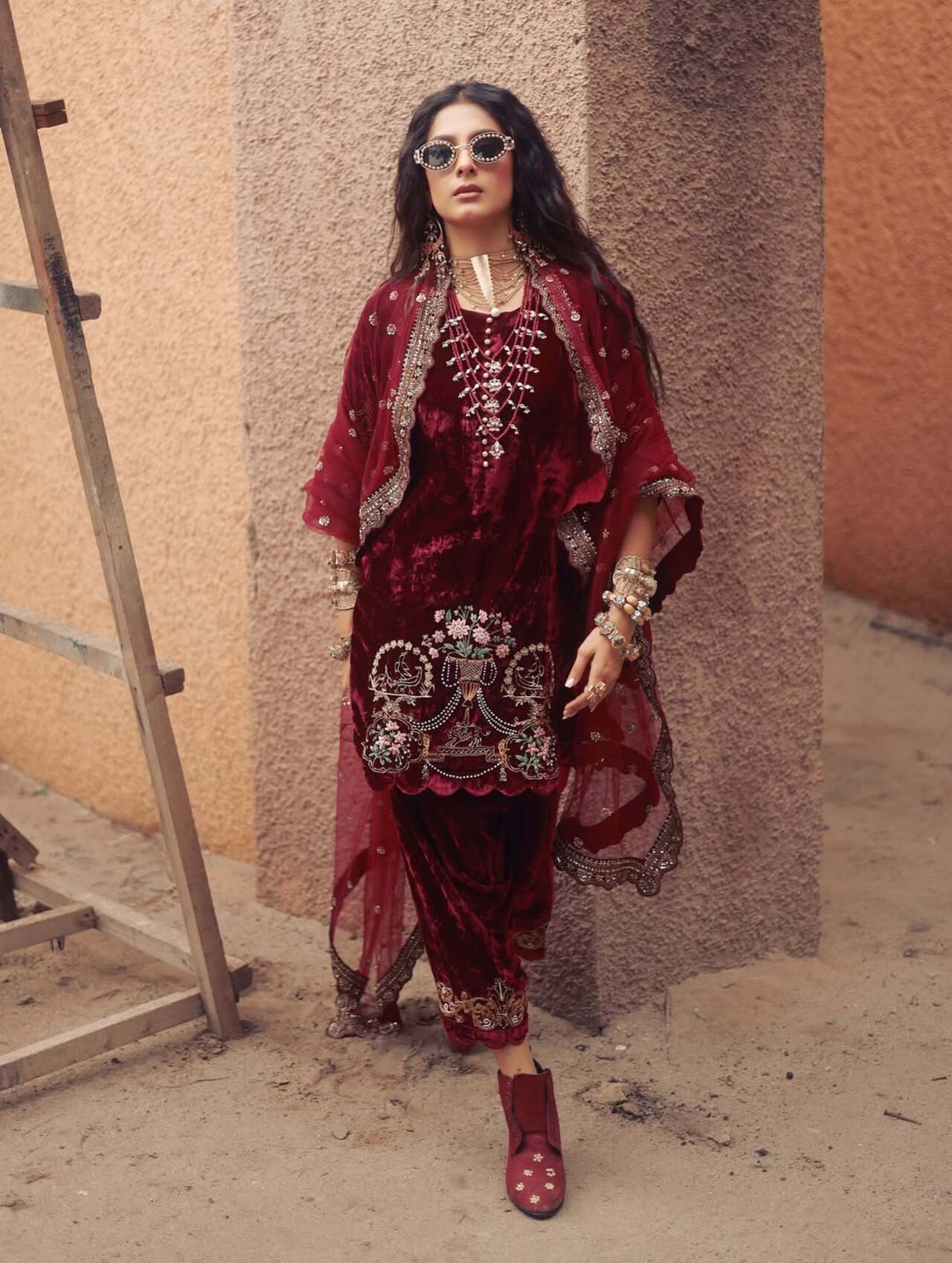 Isha Malviya is wearing a stunning maroon velvet shalwar kameez, a traditional South Asian outfit. The kameez (top) is a deep burgundy velvet with intricate gold and cream embroidery depicting a floral and baroque-inspired design centred on the front. The embroidery extends to the edges of the sleeves and hem. The shalwar (trousers) are also maroon velvet, and are a fitted, flared style that falls to the ankle