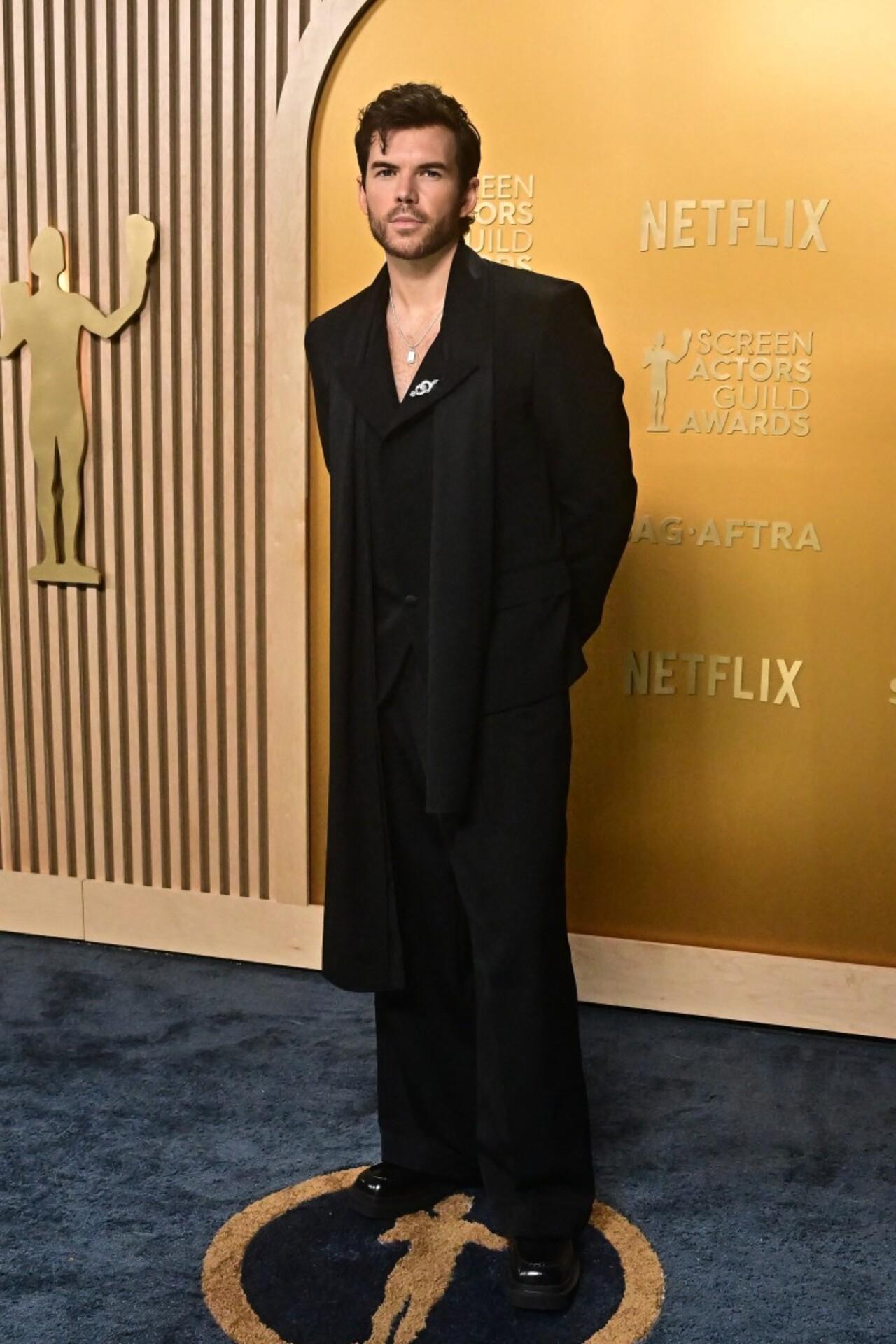 Luke Newton was dressed in a black double-breasted blazer that was layered over a black shirt with a dramatic collar and a diamond brooch pin. A long black scarf, loose-fitting pants, and black patent leather shoes completed his outfit.  