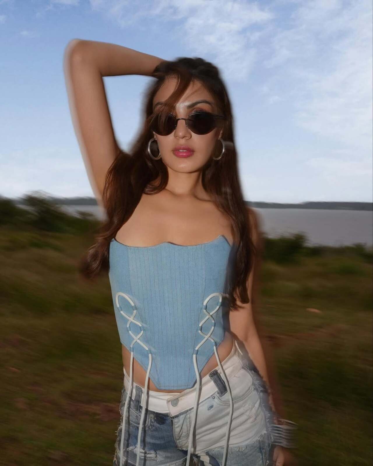 Blurred background, sharp presence. The pastel corset with lace detailing cinches her silhouette against ripped denim shorts—vacay mode, but with structure. Hoop earrings and shades add that signature pop-culture queen energy