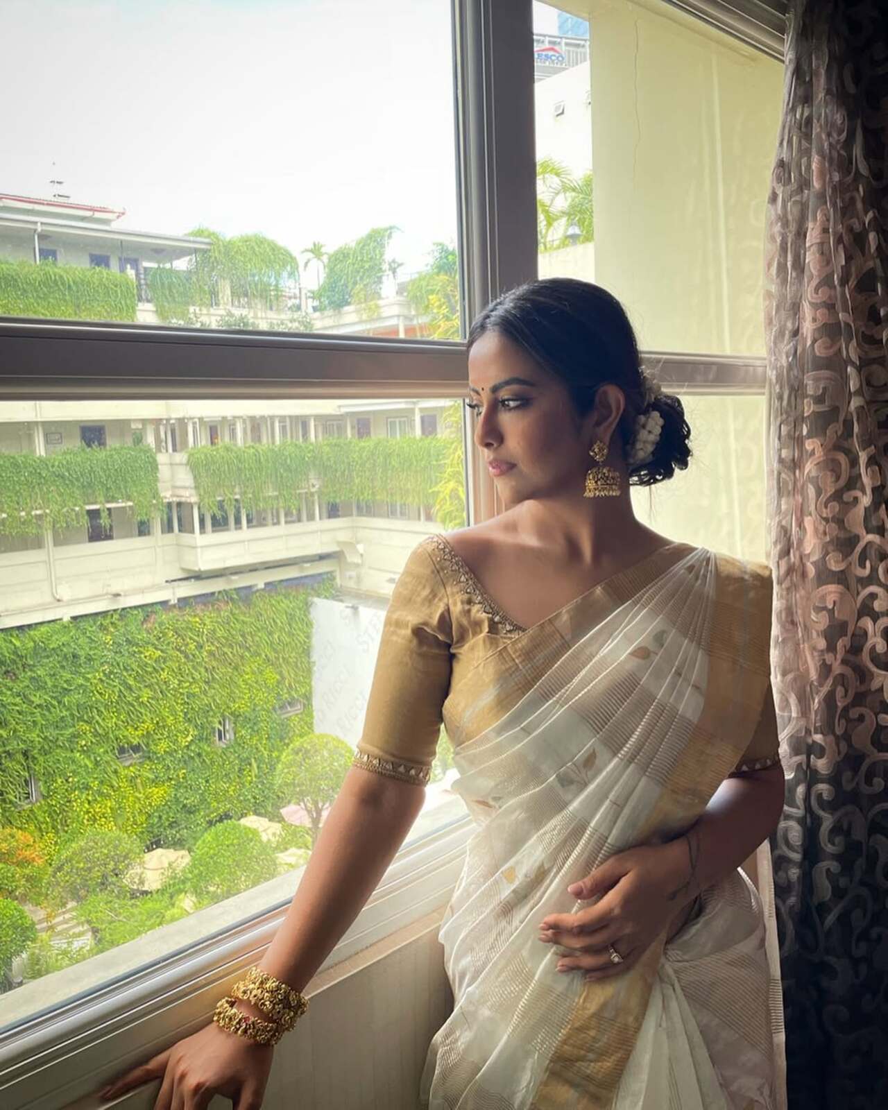 She is draped in a traditional Kerala kasavu saree, complemented by intricate jewellery. Her hair is elegantly styled into a gajra-adorned bun, while ornate jhumka baalis add a regal finishing touch