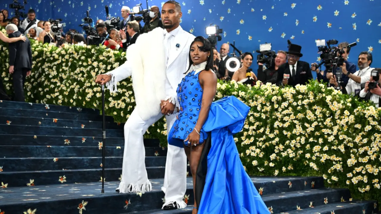 Simone Biles, the celebrated Olympic gymnast, made a dramatic entrance in a short blue dress with a sweeping train and a crisp white collar. She completed the look with sky-high heels, exuding both strength and style. Her husband, Jonathan Owens, safety for the Chicago Bears, wore a striking white suit accessorized with a fur-draped shoulder and a stylish cane