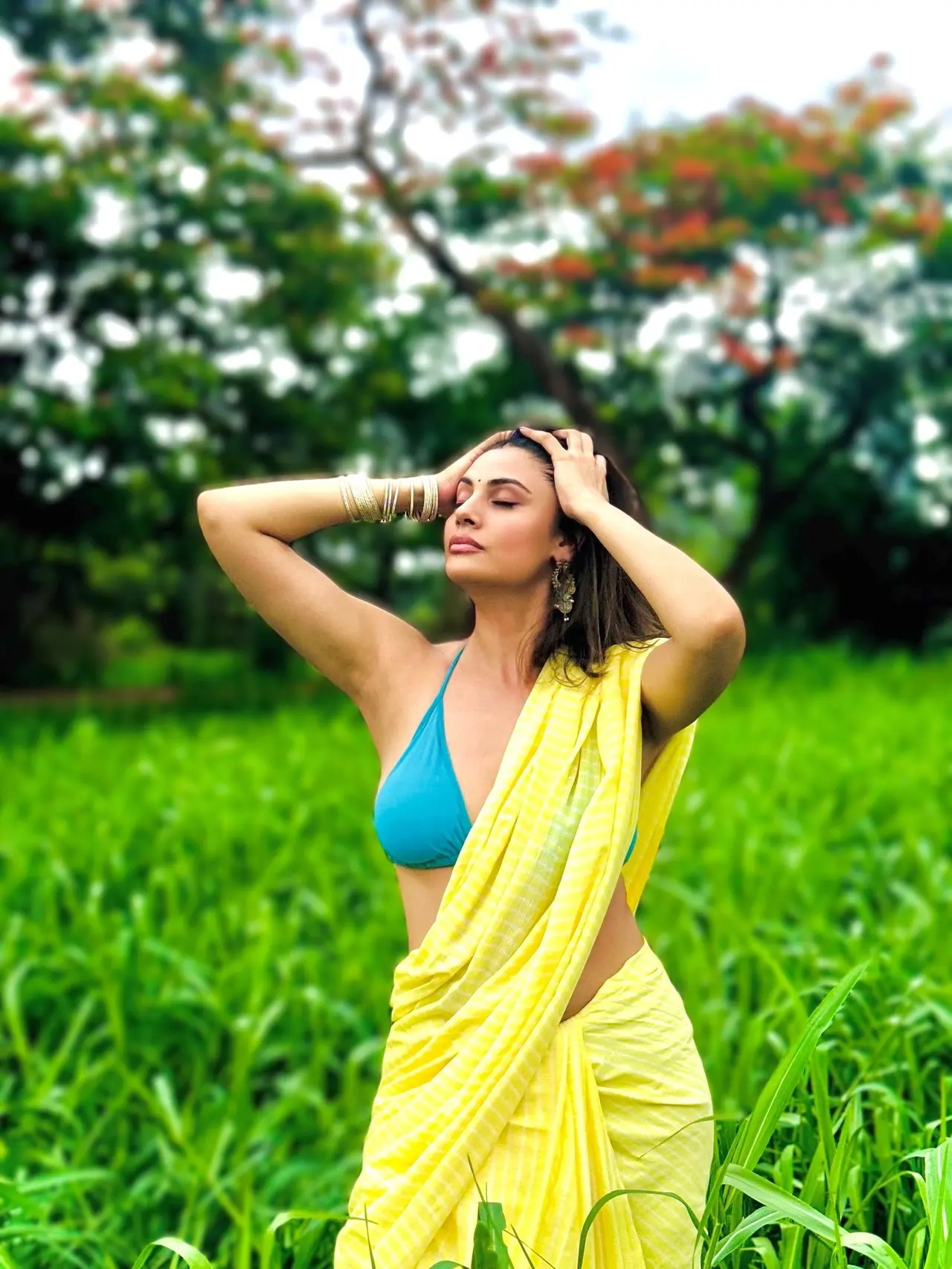 Raising the temperatures in this yellow saree, Malti pairs it with a sultry blue bralette while posing in the fields. She completes the look with hanging earrings and golden bangles.