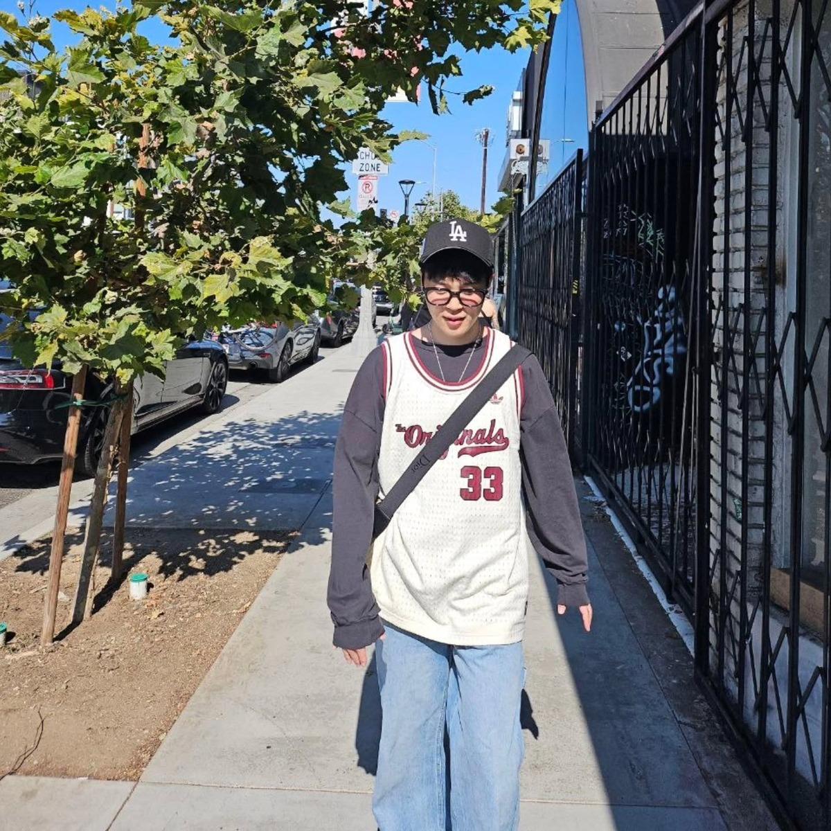 Chanelling his inner nerd, BTS Jimin pairs a jersey vest with a long-sleeved t-shirt and blue jeans. He completes the look with a cross-body bag and a cap.
