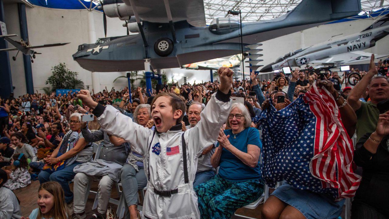 A young boy in an astronaut costume cheers beside a woman waving a flag as they watch the live broadcast of the Artemis II crew&rsquo;s splashdown at the San Diego Air and Space Museum, celebrating the astronauts&rsquo; safe return
