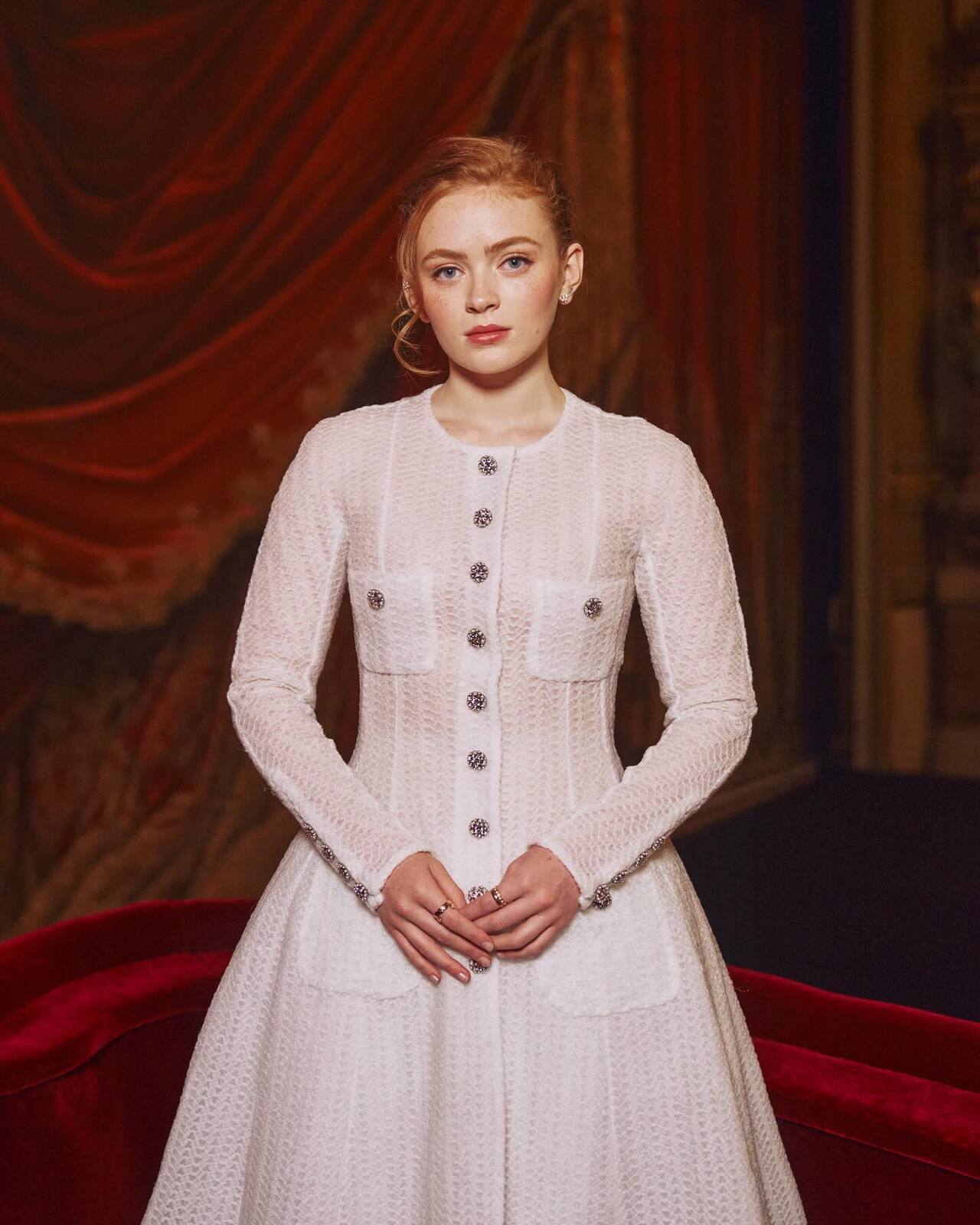 Sadie Sink looks classic in this ivory textured A-line dress, featuring long sleeves and a sophisticated button-down front. The garment is detailed with shimmering jeweled buttons.