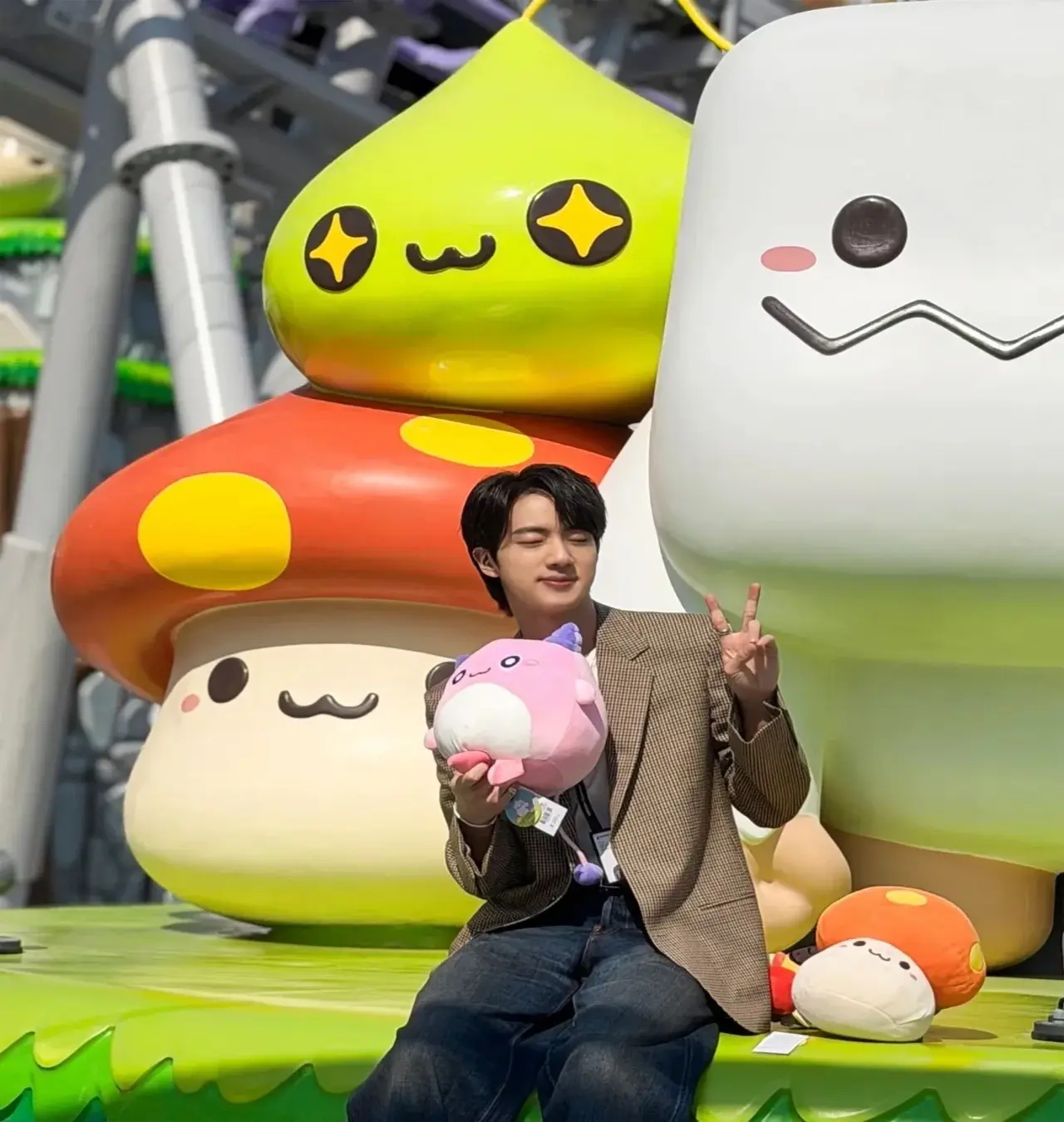 Jin's love for Maple Story is well documented. Here he poses happily behind with a giant Orange Mushroom statue at the back. He holds a Pink Bean mushroom plushie in hand