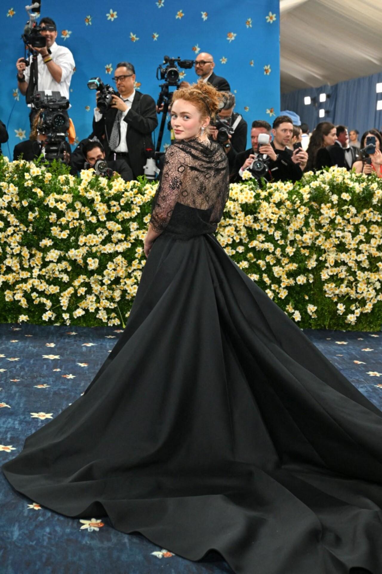 Sadie Sink brings a dark, romantic energy to the Met Gala in this monochrome black Alexander McQueen ensemble. The look features a floor-sweeping satin skirt paired with a delicate sheer lace top adorned with intricate floral embroidery.