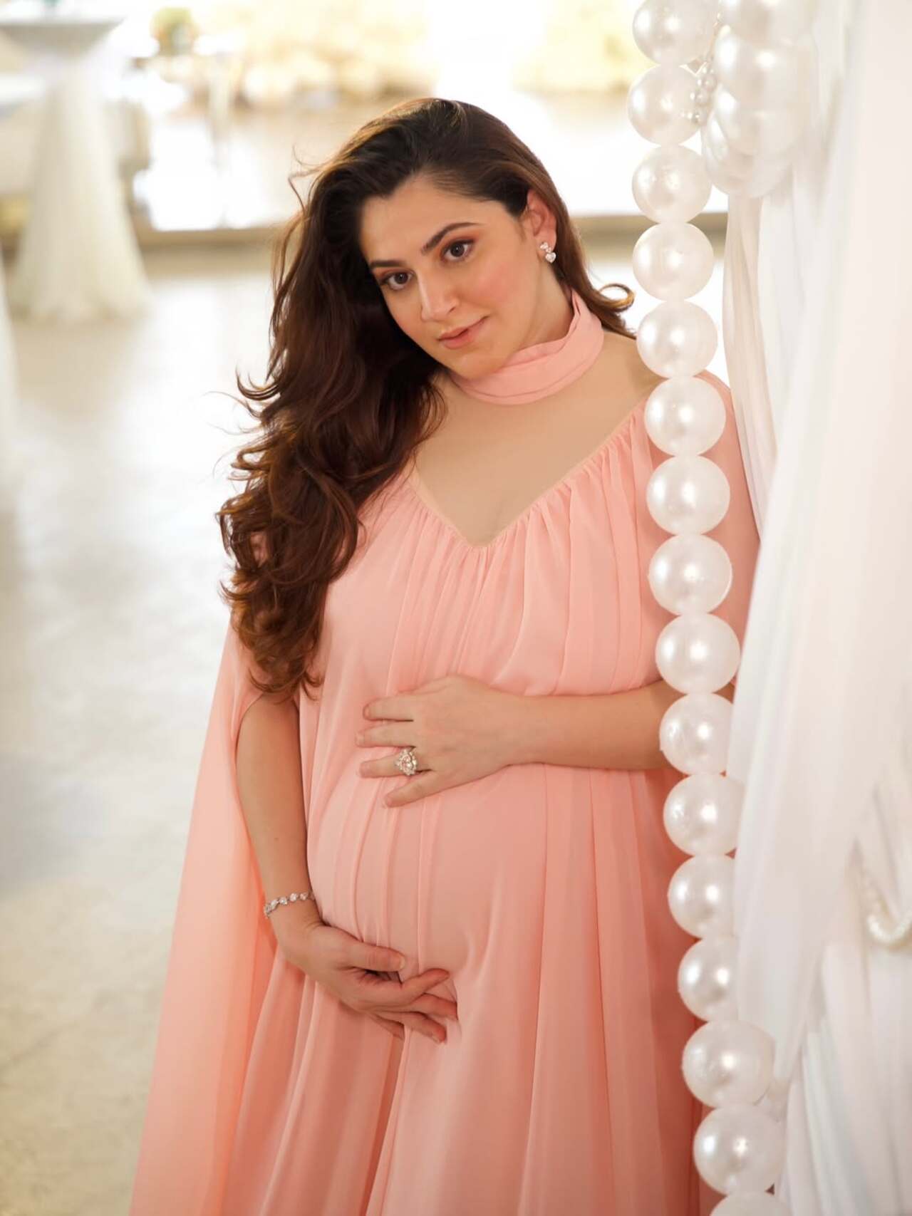 Shivaleeka Oberoi exudes soft elegance in a pastel peach flowy maternity gown with delicate pleats and cape-style sleeves. While minimal jewellery and dewy makeup enhance her natural glow.