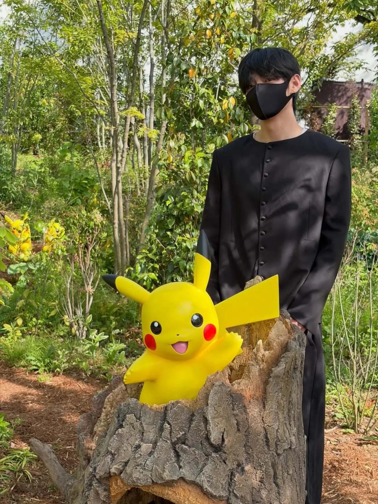 Dressed in a sleek all-black outfit with a matching face mask, he stands beside a cheerful Pikachu figure popping out of a tree stump.&nbsp;