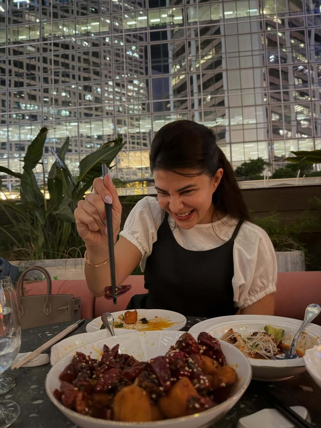 Jacqueline Fernandez is seen enjoying a lively dining moment, smiling as she picks up food with chopsticks. Surrounded by a spread of dishes, she looks completely immersed in the experience.