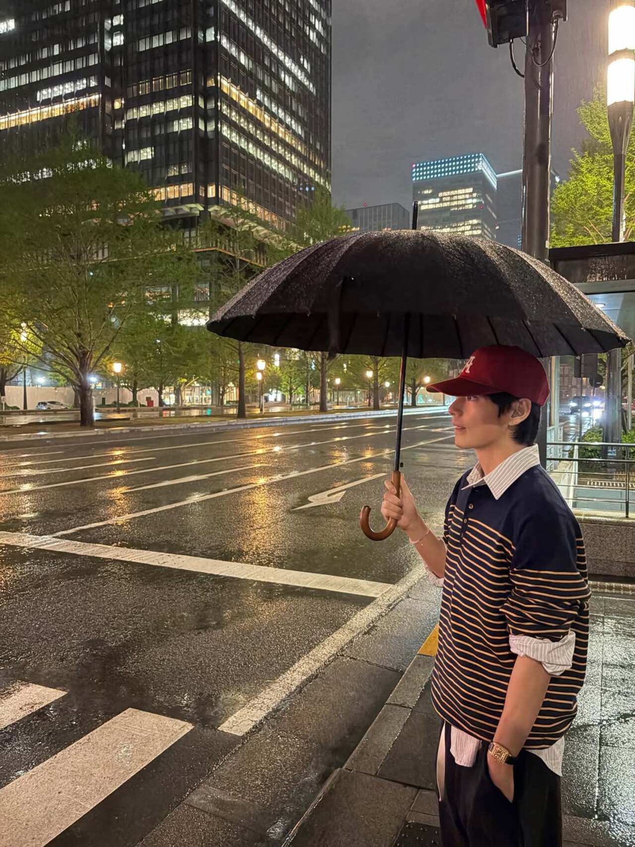 V is seen enjoying a serene moment on the streets of Japan on a rainy night. Holding a black umbrella, he stands by a quiet, glistening road soaking in the calm, late-night urban vibe during his time in Japan.