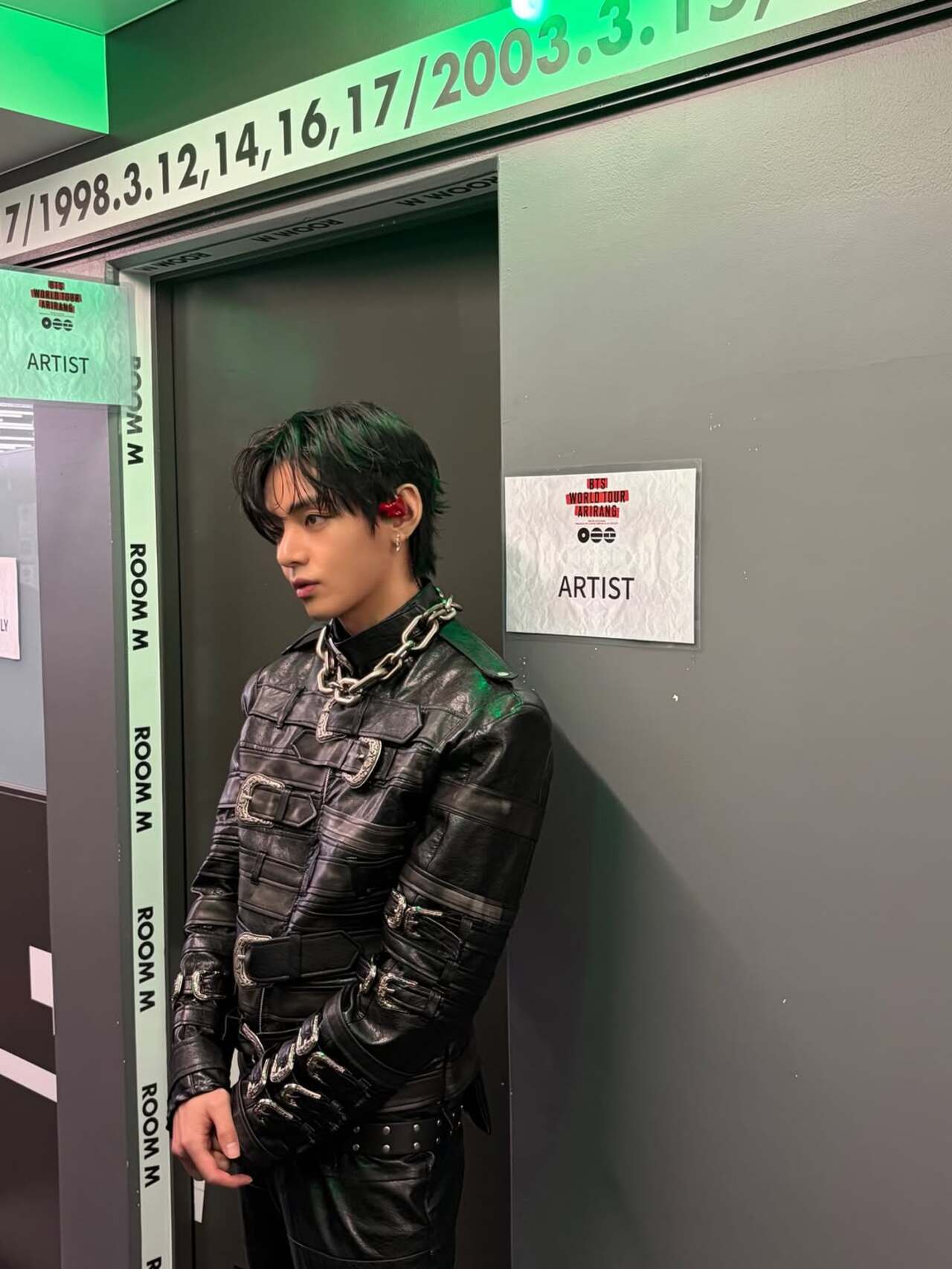V is seen at backstage in a striking performance look, dressed in an all-black leather outfit detailed with buckles, straps, and metallic accents.