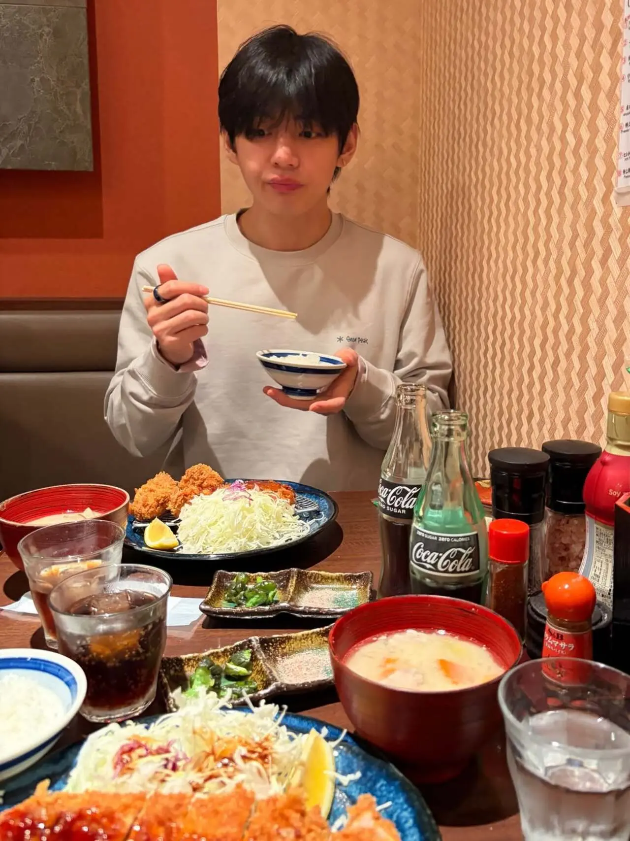 Kim Taehyung is seen enjoying a wholesome Japanese meal at a cozy restaurant. He holds a bowl of rice with chopsticks, while his table is filled with dishes like crispy tonkatsu (breaded pork cutlet), finely shredded cabbage salad, a bowl of warm miso soup.