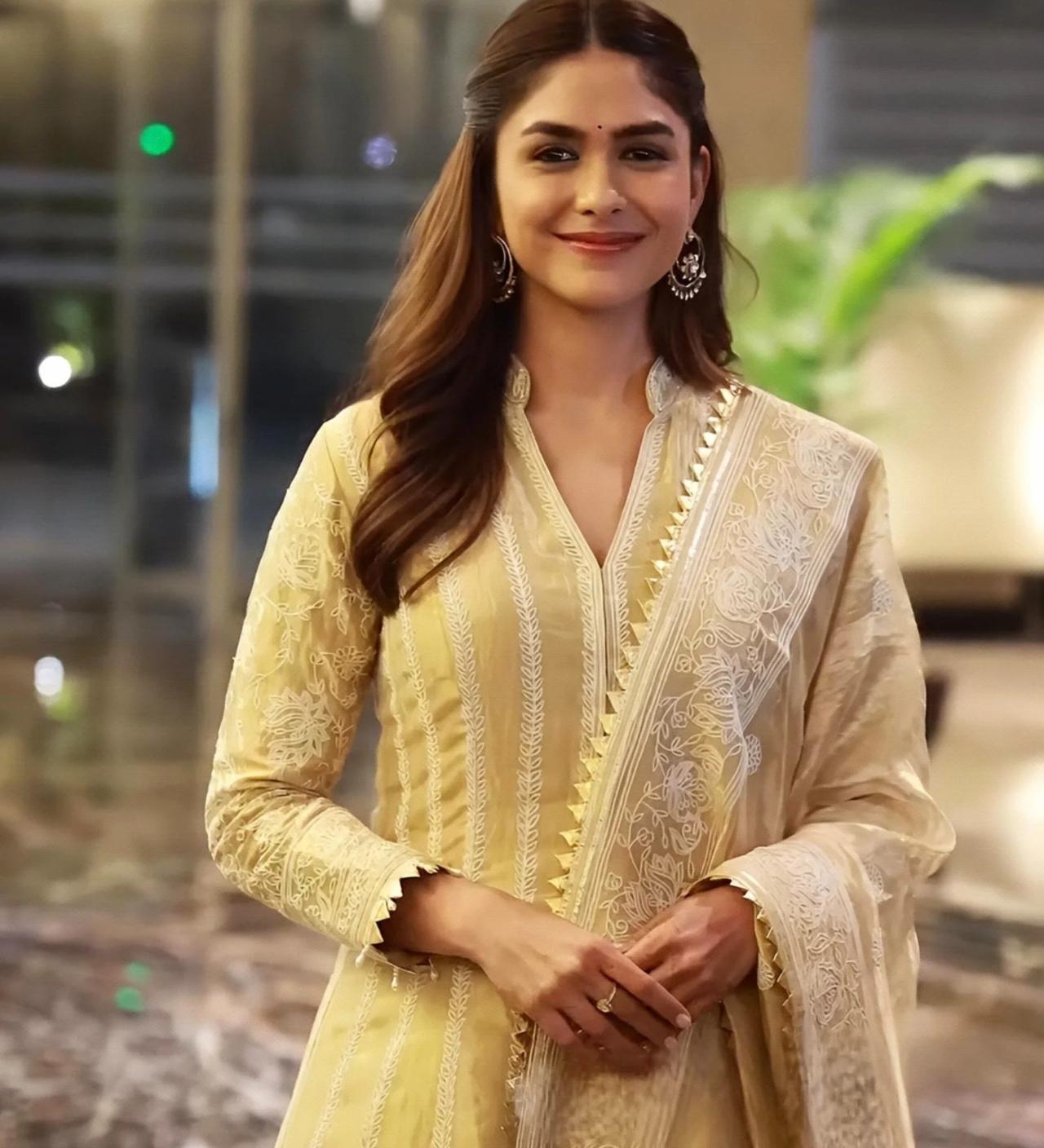 Mrunal Thakur embraces a timeless, ethnic aesthetic in this radiant pale yellow Anarkali suit. The ensemble features intricate delicate lace detailing and is paired with a matching sheer dupatta that adds a layer of ethereal grace.