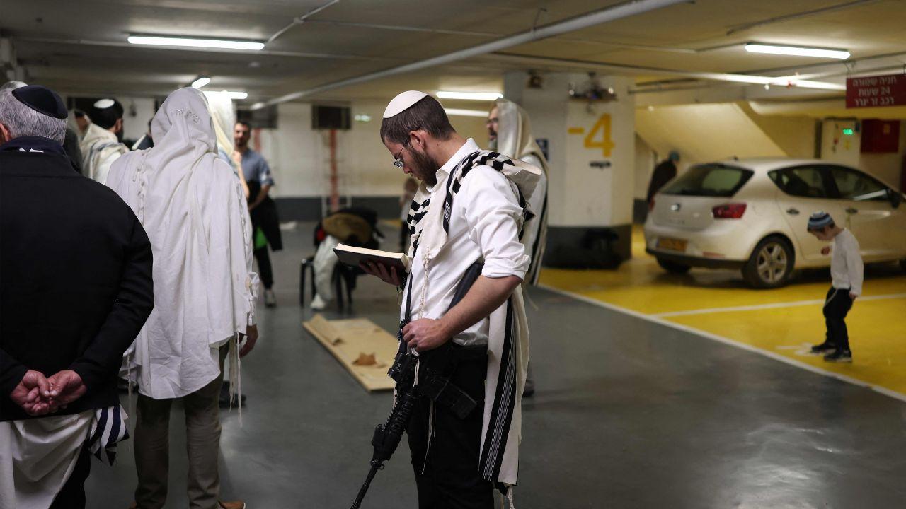 An armed man prays while civilians take cover in Tel Aviv, blending routine religious observance with emergency preparedness