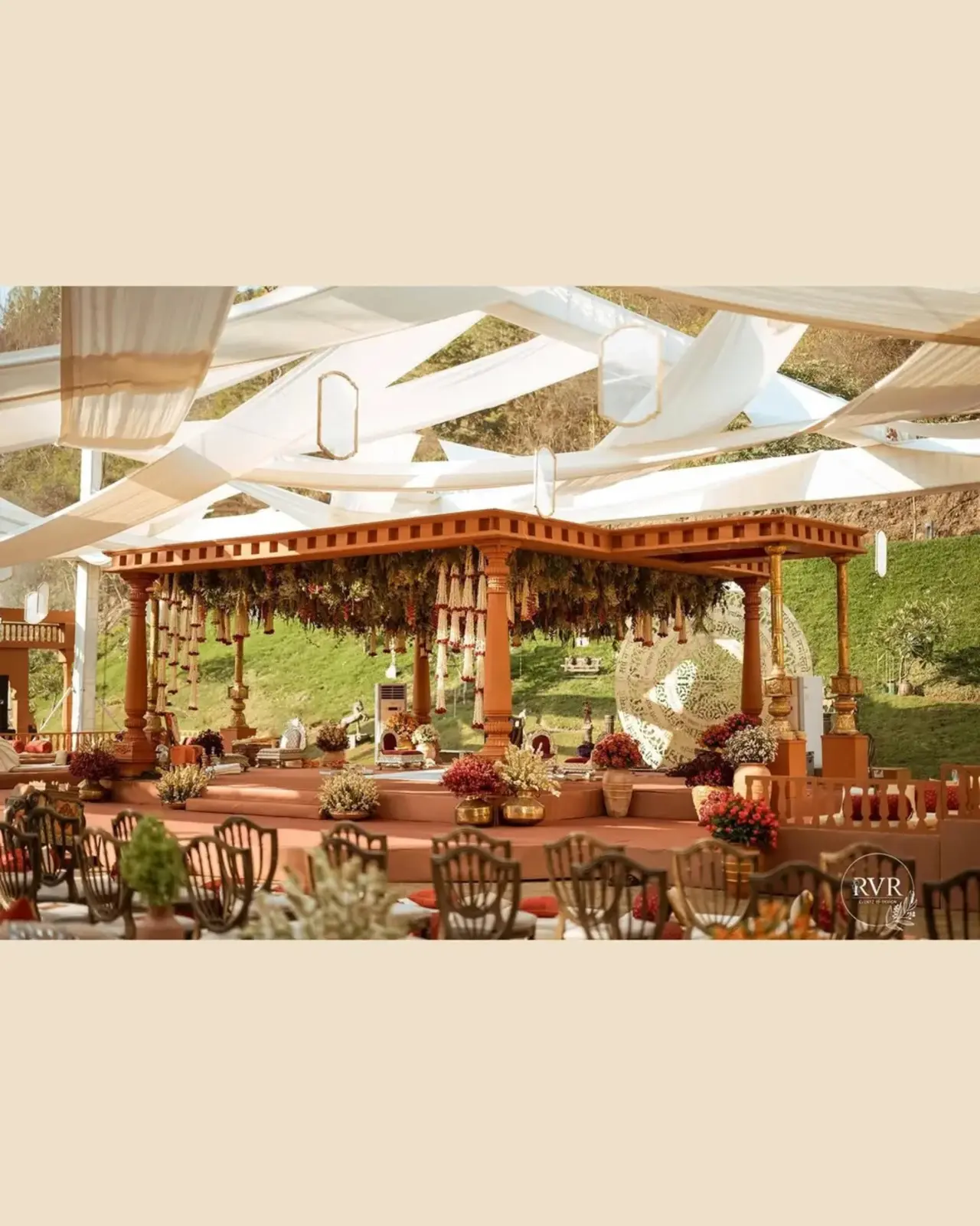 The color palette is rich and warm dominated by terracotta, rust, deep maroon and antique gold. The mandap area features intricately carved wooden pillars and traditional architectural elements reminiscent of temple design