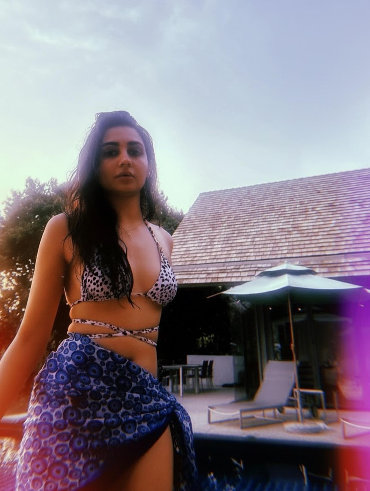 Vedika wears a white-and-black printed halter bikini top paired with a blue printed sarong tied at the waist. Her loose, wet hair and minimal accessories complement the tropical villa setting, creating a relaxed vacation look.