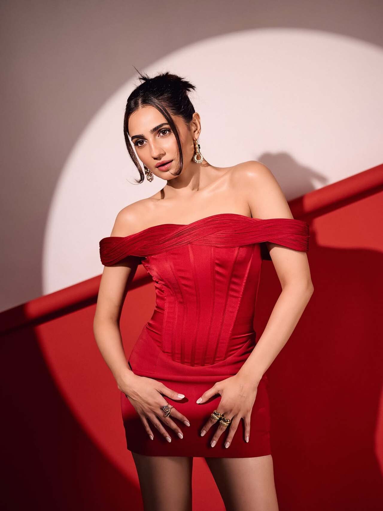 She wears an off-shoulder structured red mini dress with corset-inspired detailing. A sleek updo, drop earrings and stacked rings complete the vibrant, high-fashion look against the matching red backdrop