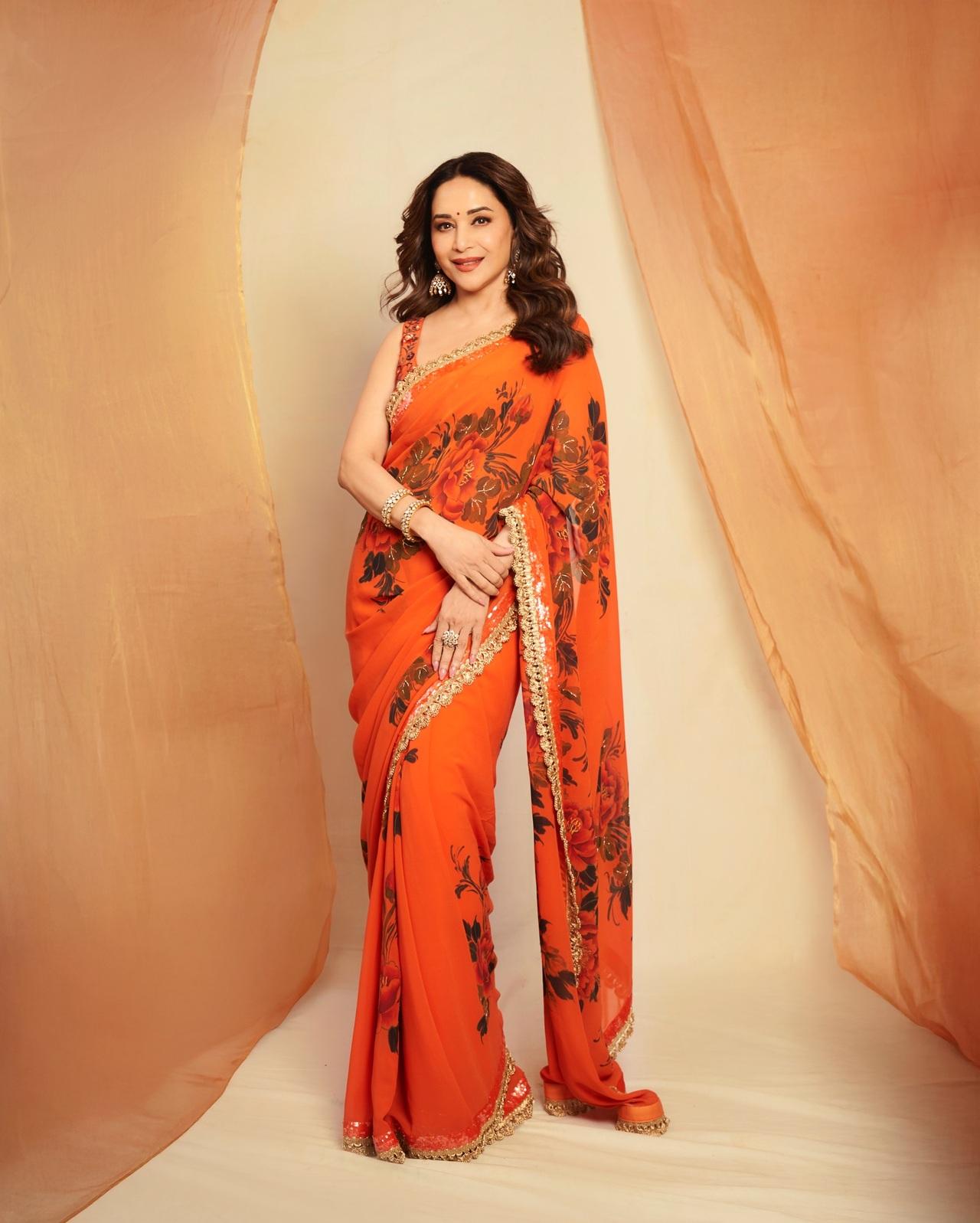 Madhuri Dixit looks absolutely stunning and elegant in a vibrant orange saree adorned with beautiful floral prints and a shimmering golden border.