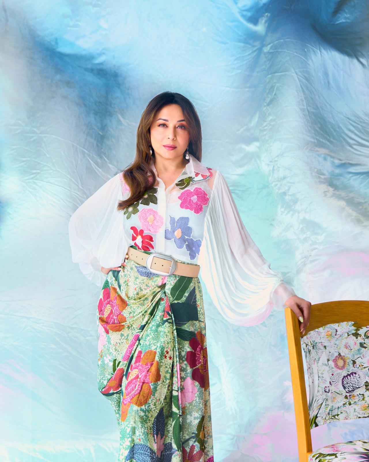 Madhuri Dixit radiates effortless charm in a chic Indo-western ensemble, featuring a semi-sheer white shirt with voluminous statement sleeves and bright, colorful floral appliques. She pairs the top with a draped, wrap-style green skirt adorned in matching vibrant floral prints.