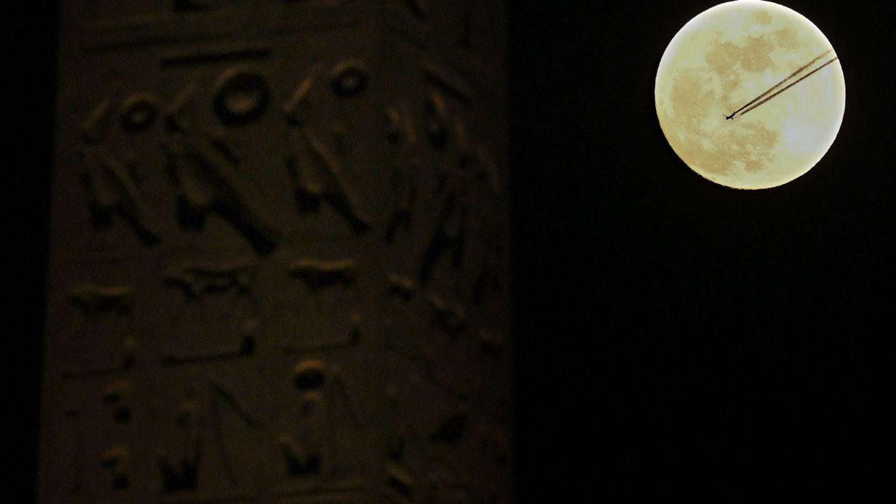 An aircraft cuts across the sky beside the full moon framed by Luxor Obelisks at Place de la Concorde