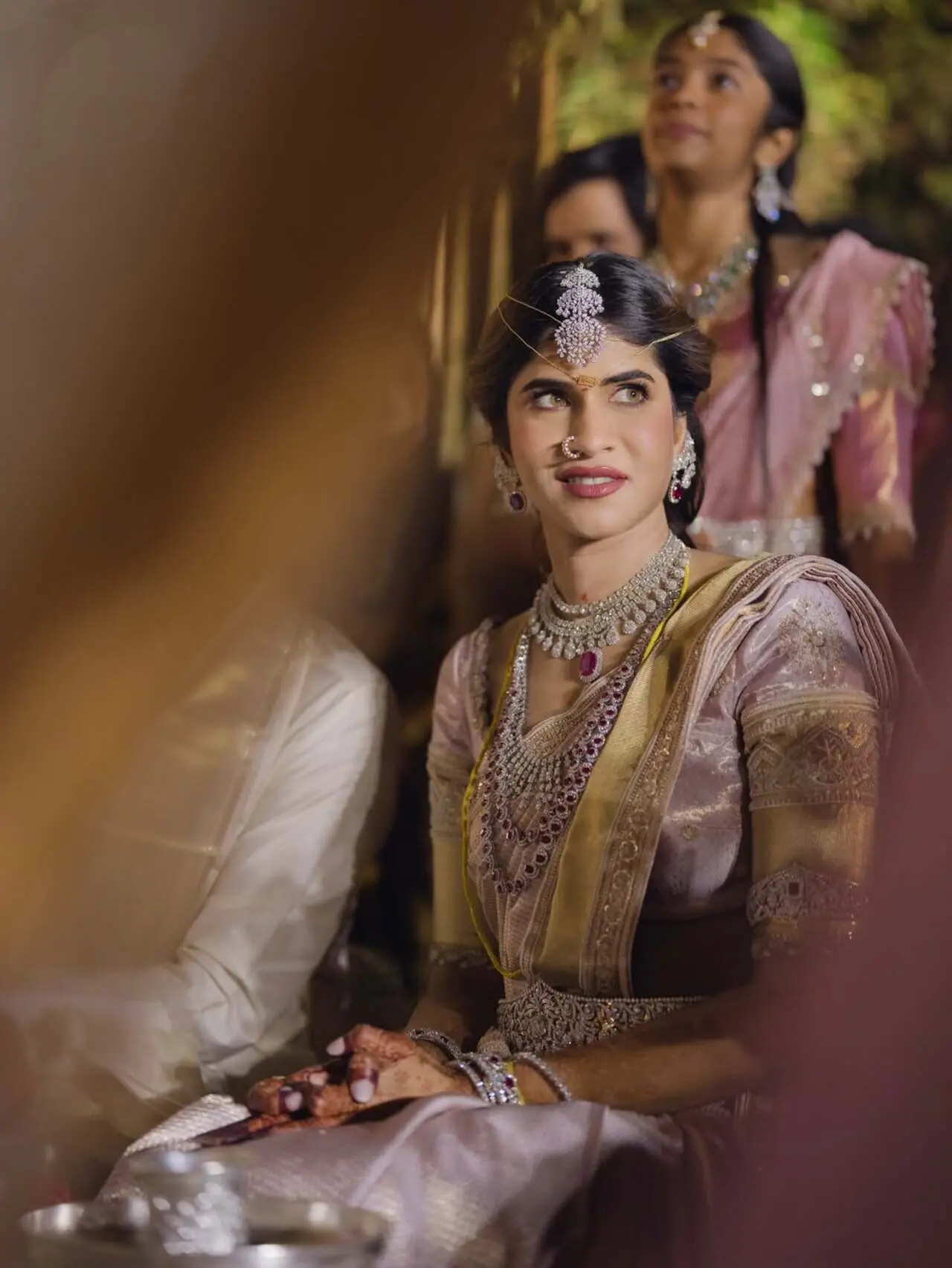 Nayanika is dressed in a soft pink silk saree with rich gold borders, making her look very graceful and festive. She is wearing layered necklaces, earrings, bangles, and a delicate headpiece that make her look stunning.