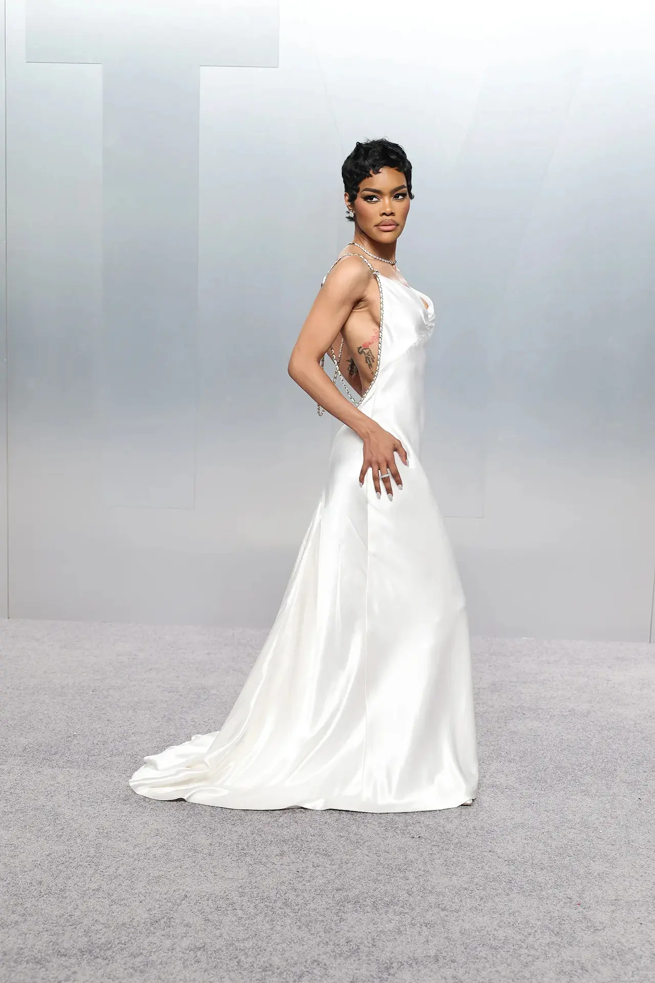 Teyana Taylor stunned in a white satin dress that fell in a long, narrow silhouette. The gown featured a softly scooped neckline with a lightly gathered bust, giving the front a slip-dress effect. The sides and back opened dramatically beneath chain-trimmed straps, adding a bold edge.