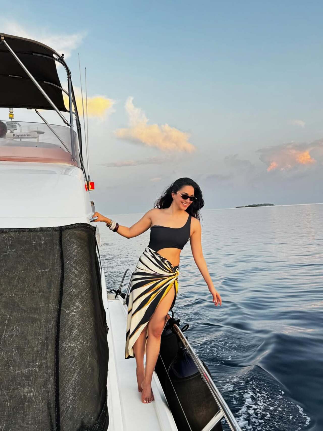 Kiara Advani looks effortlessly elegant as she poses on a yacht against a serene ocean backdrop. Dressed in a chic one-shoulder black top paired with a flowing, patterned sarong, she exudes a perfect blend of glamour and ease.